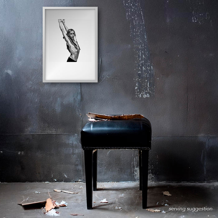 Framed B&W portrait of Iggy Pop by Gavin Evans, 1996. Profile pose, arms skyward, intense gaze, feathered crown. Displayed in raw interior, Iggy Pop Collection.